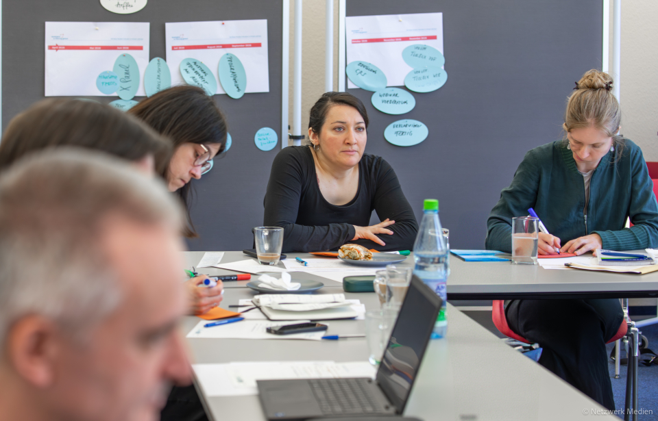 5 Personen sitzen an einem Tisch während einer Arbeitsphase. 1 Frau blickt nach vorne, die anderen Personen blicken nach und machen Notizen.