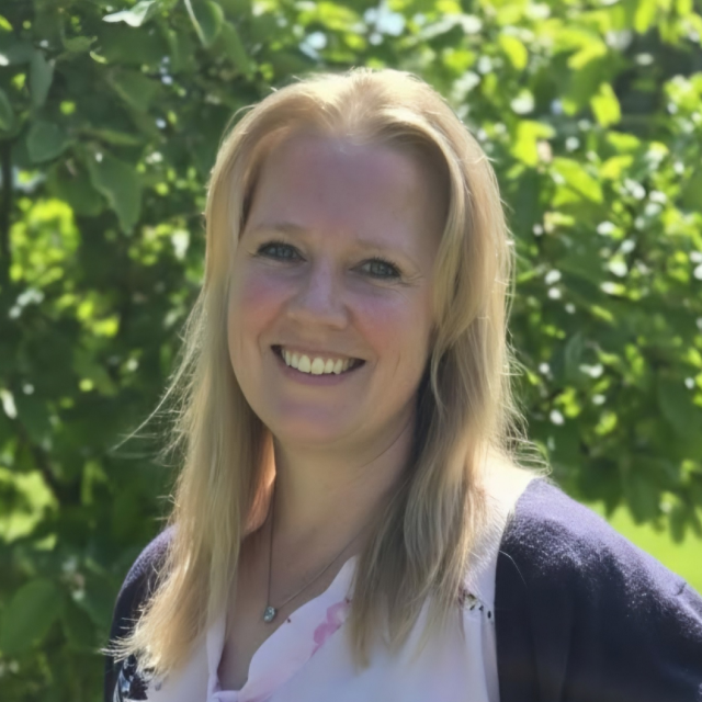 Porträt von Verena Gronninger. Sie hat schulterlanges, blondes Haar und trägt ein dunkelblaues Oberteil und ein buntes Halstuch mit Blumenmuster.