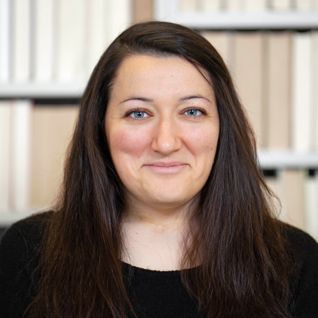 Porträt von Rebecca Groß. Sie hat langes, dunkles Haar trägt ein schwarzes Oberteil. Im Hintergrund ist ein unscharfer Innenraum mit hellen Wänden. Porträt von Rebecca Groß. Sie hat langes, dunkles Haar trägt ein schwarzes Oberteil. Im Hintergrund ist ein unscharfer Innenraum mit hellen Wänden.