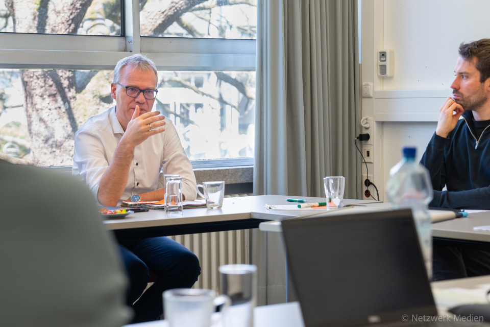 1 Mann mit Brille sitzt am Tisch und gestikuliert mit der Hand, während er etwas erklärt. 1 Mann rechts sitzend hört zu. 