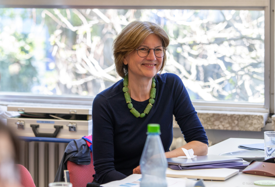 Frau Prof. Niehaus sitzt an einem Tisch und lächelt. Sie trägt einen blauen Pullover mit einer grünen Halskette.