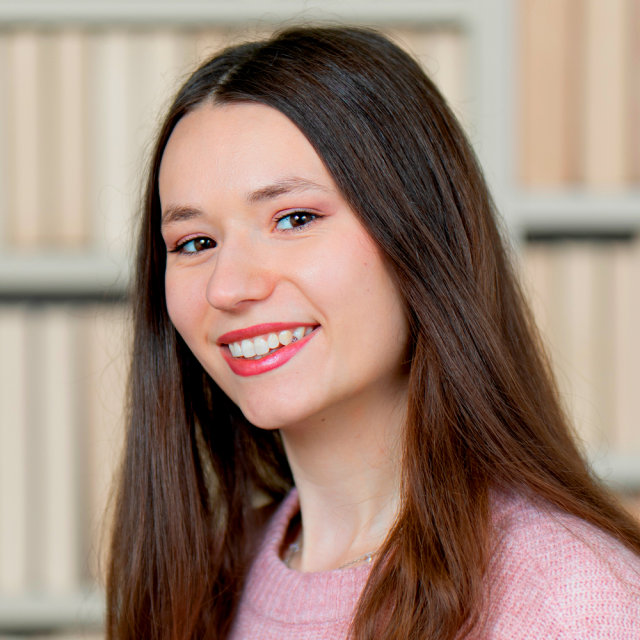 Porträt von Melissa Kruft. Sie hat langes braunes Haar und trägt einen rosa Pullover. Im Hintergrund ist ein unscharfer Innenraum mit hellen Wänden. Porträt von Melissa Kruft. Sie hat langes braunes Haar und trägt einen rosa Pullover. Im Hintergrund ist ein unscharfer Innenraum mit hellen Wänden.