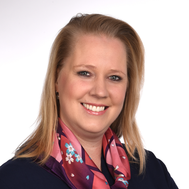 Porträt von Verena Gronninger. Sie hat schulterlanges, blondes Haar und trägt ein dunkelblaues Oberteil und ein buntes Halstuch mit Blumenmuster.