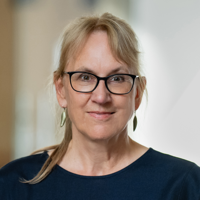 Porträt von Helga Albrecht. Sie trägt eine Brille, einen Zopf und hat blondes Haar. Das Oberteil ist dunkelblau. Der Hintergrund ist unscharf.